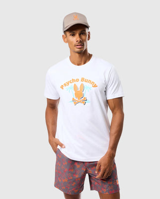 A man is wearing the "MENS BOSTON GRAPHIC TEE - B6U573C200" by Psycho Bunny, a white t-shirt crafted from Peruvian Pima cotton featuring an orange bunny logo printed on the front. He pairs it with a beige cap and gray shorts adorned with red bunny patterns. The background is plain light gray.