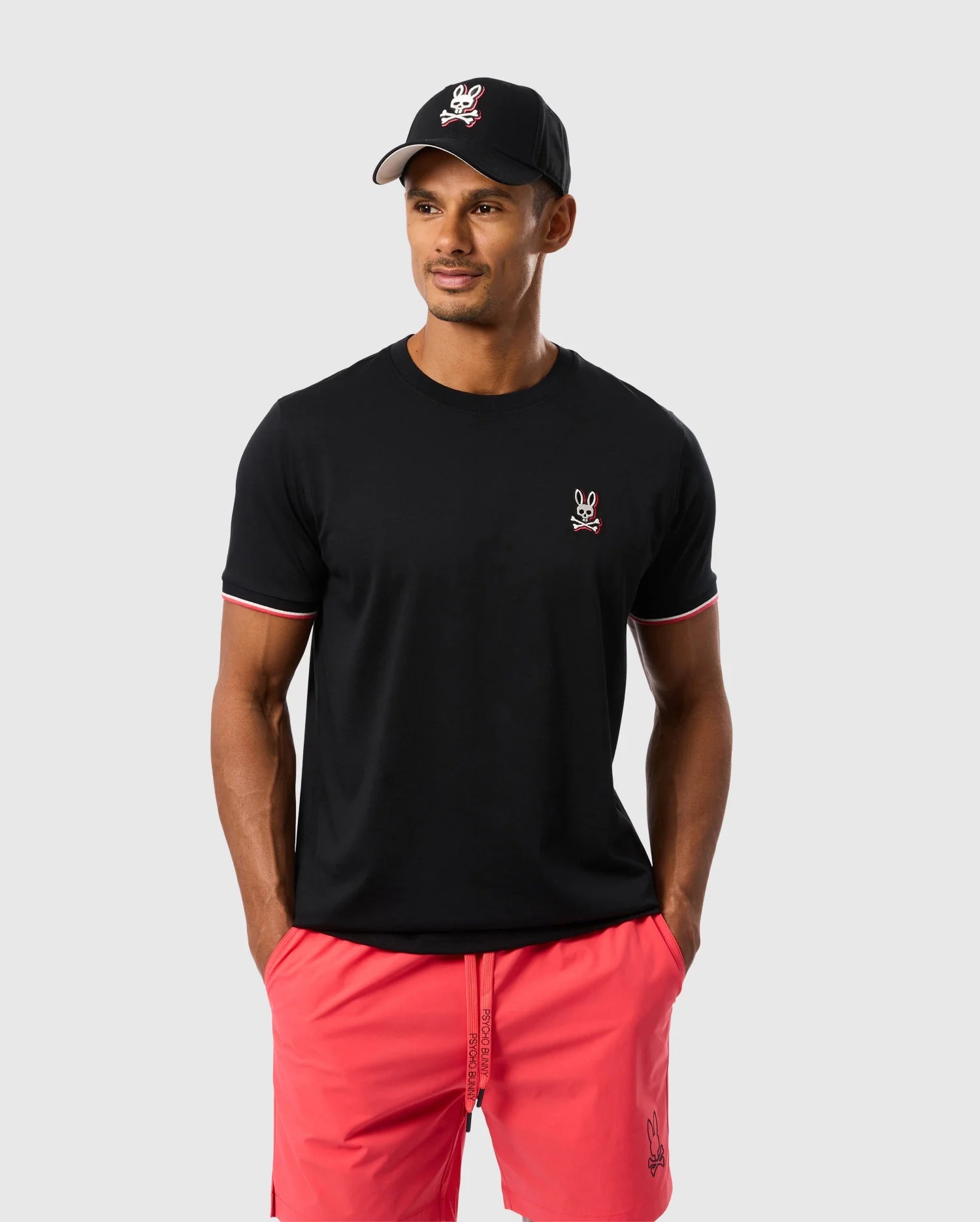 A man is wearing the Psycho Bunny MENS KAYDEN FASHION TEE - B6U577C200, a black tee made of Pima cotton with a small embroidered rabbit emblem. He pairs it with red shorts and a black baseball cap featuring a similar rabbit logo. He stands against a plain gray background, looking slightly to his left with his hands in his shorts pockets.