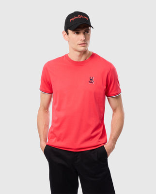 A man stands against a plain background, wearing a vibrant red Psycho Bunny MENS KAYDEN FASHION TEE - B6U577C200, crafted from soft Pima cotton and adorned with a small embroidered logo on the chest. He pairs it with black pants and a black baseball cap featuring red lettering. His hands are in his pockets as he gazes slightly to the side.