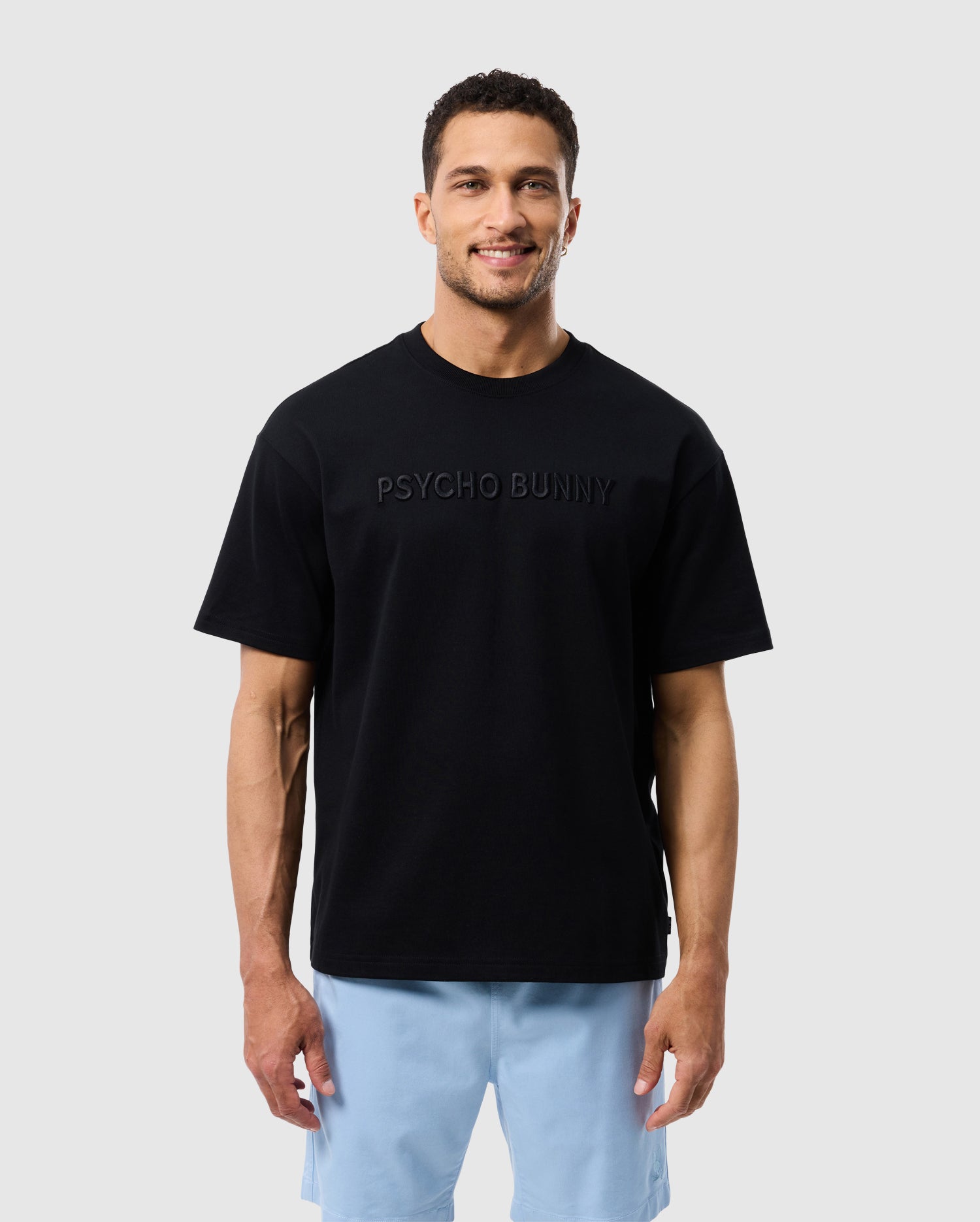 A man with short, dark curly hair stands against a plain background, smiling. He is wearing the Psycho Bunny MENS WINDCREST HEAVY WEIGHT TEE - B6U593C200 in black and light blue shorts.