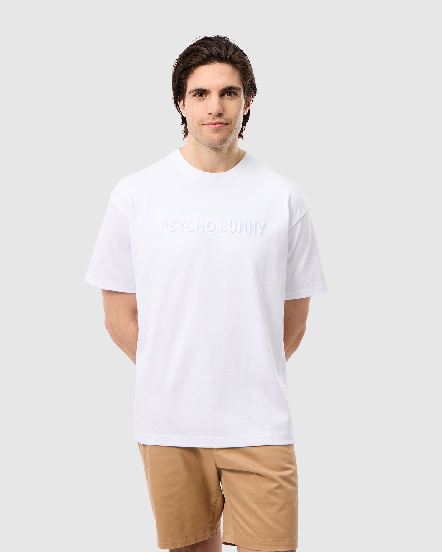 A man with dark hair is wearing the Psycho Bunny Men's Windcrest Heavy Weight Tee (B6U593C200), a relaxed fit white Pima cotton shirt featuring the text 