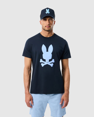 A man is wearing the MENS HOUSTON GRAPHIC TEE - B6U607C200 by Psycho Bunny, a navy soft Pima cotton shirt featuring their iconic white bunny skull and crossbones graphic. He pairs it with light blue shorts against a plain background.