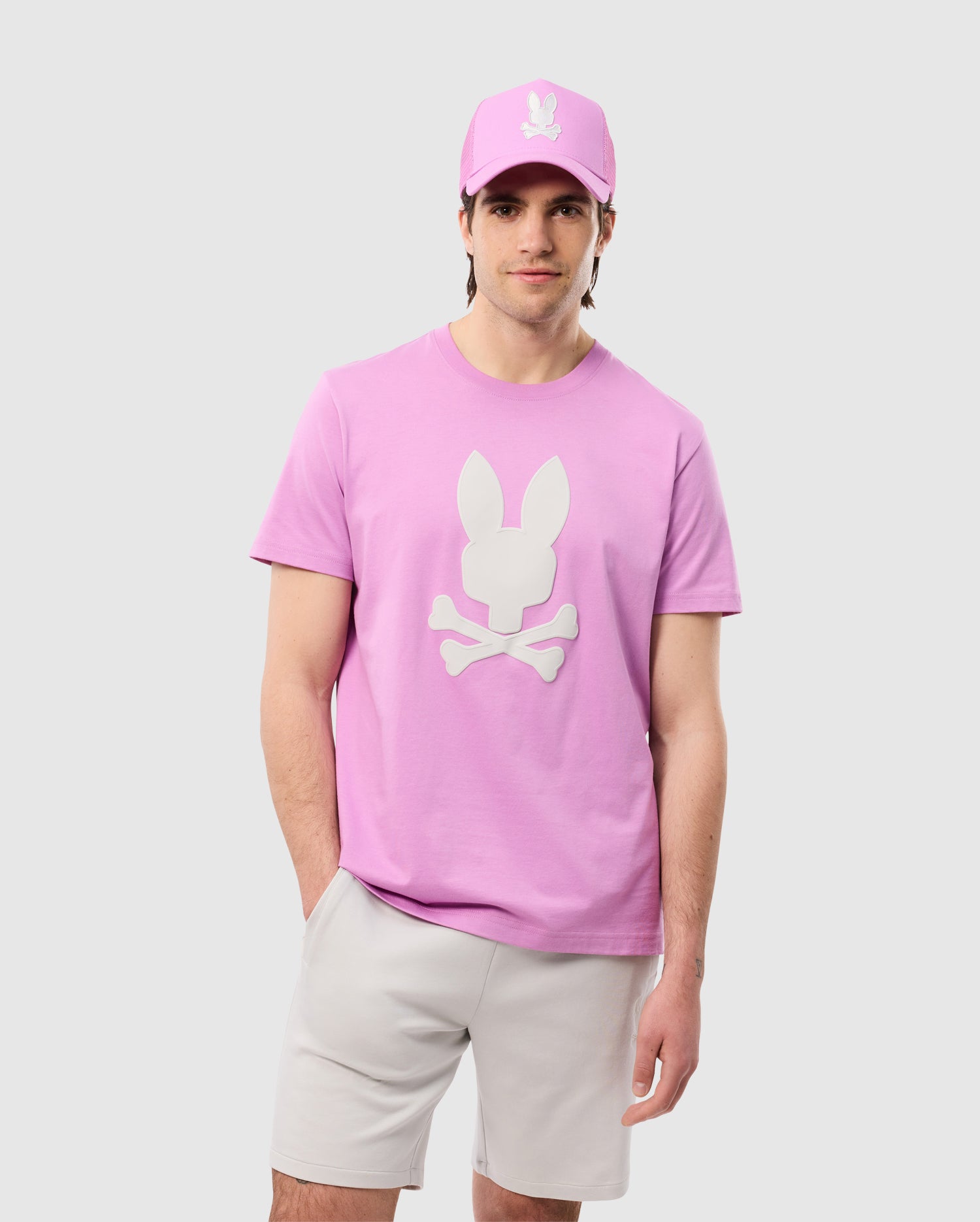 A man stands against a plain background wearing a pink Psycho Bunny MENS HOUSTON GRAPHIC TEE - B6U607C200 with a white bunny and crossbones graphic, made from soft Pima cotton. He pairs it with a matching pink cap and light gray shorts. With one hand in his pocket, he gazes directly at the camera with a slight smile.