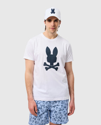 A man wearing a white Psycho Bunny MENS HOUSTON GRAPHIC TEE - B6U607C200 with a black bunny and crossbones logo, a matching white baseball cap, and blue shorts featuring a playful Bunny print stands against a plain, light grey background.