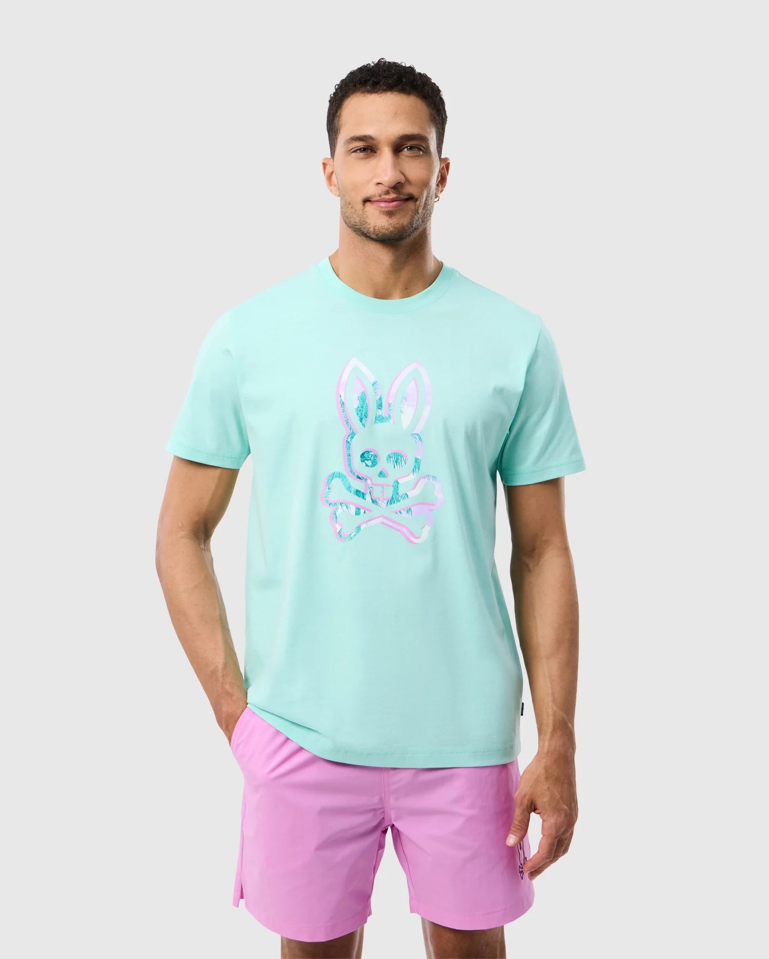 A man in a light blue Psycho Bunny MENS LEONARD GRAPHIC TEE - B6U609C200 featuring a pink bunny skull design stands against a plain background. He is also wearing pink shorts and has a short haircut. Made from soft Pima cotton, he looks straight at the camera with a slight smile.