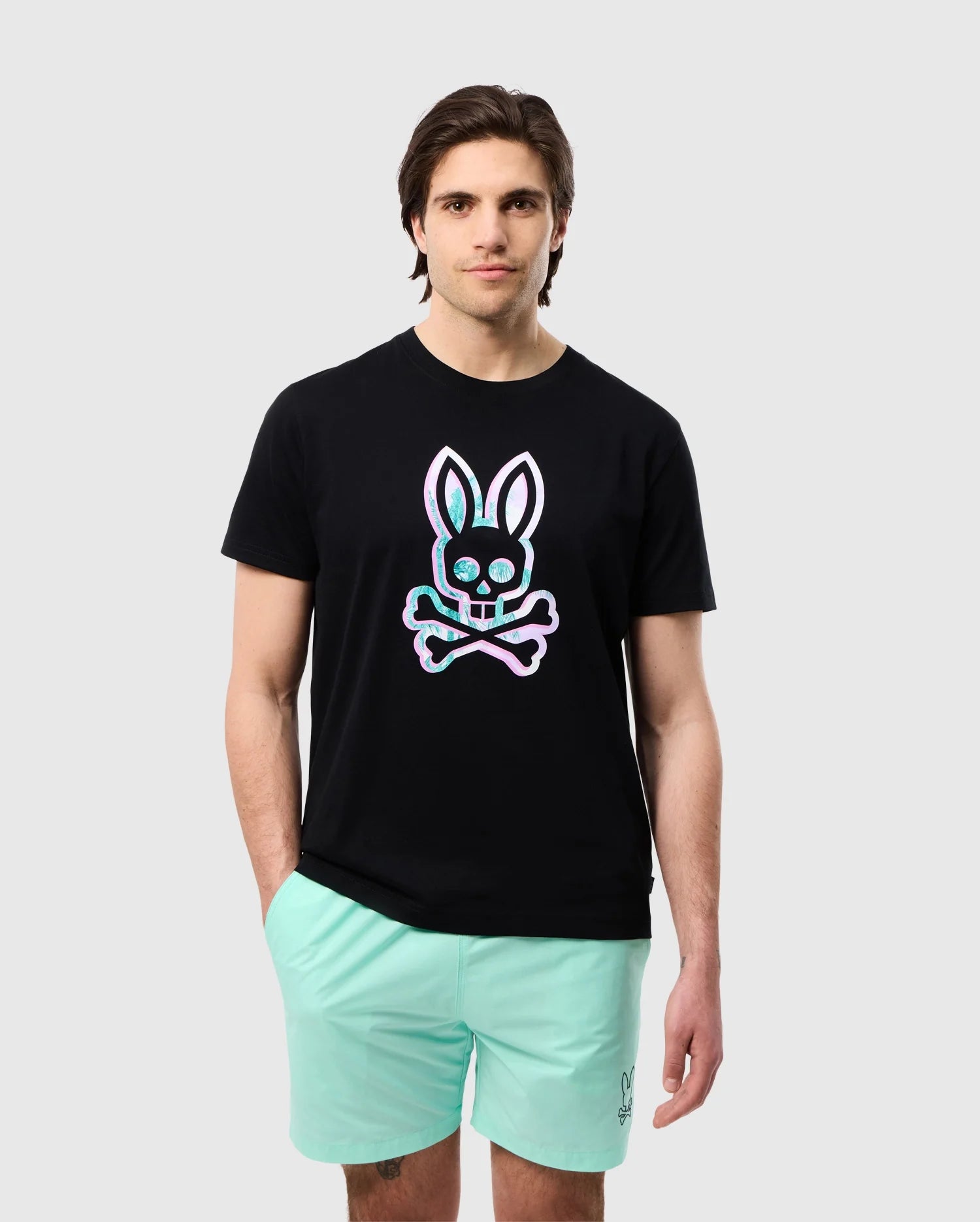 A man with medium-length brown hair is wearing a high-quality Psycho Bunny MENS LEONARD GRAPHIC TEE - B6U609C200 featuring a colorful skull and crossbones with bunny ears graphic. Made from luxurious Pima cotton, the black T-shirt pairs perfectly with his mint green shorts against the plain light gray background.