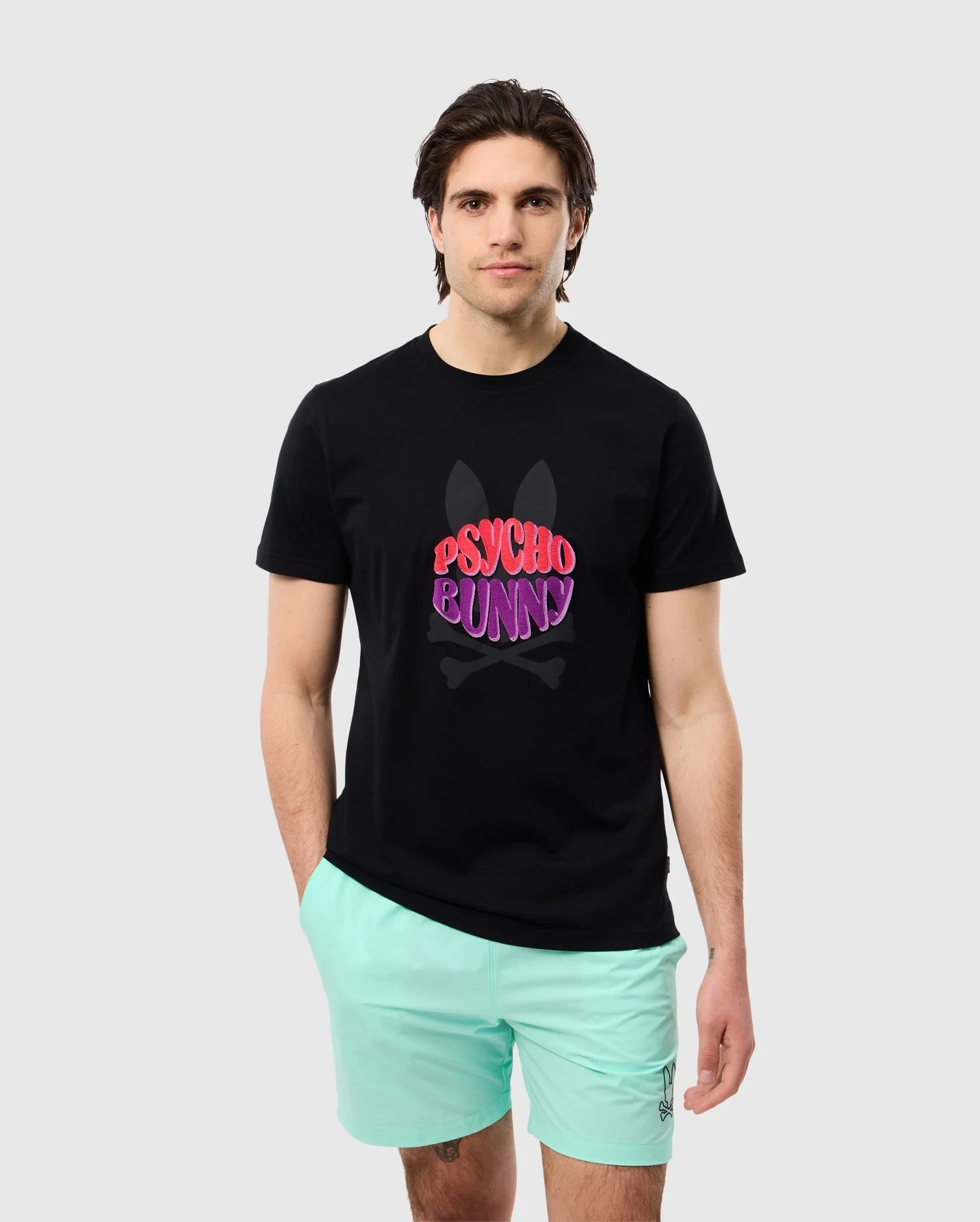 A person with dark hair wearing a black MENS PRESTON GRAPHIC TEE - B6U611C200 by Psycho Bunny and mint green shorts stands against a plain light grey background. The high-quality Pima cotton t-shirt features a colorful logo with the brand name over a bunny skull design.