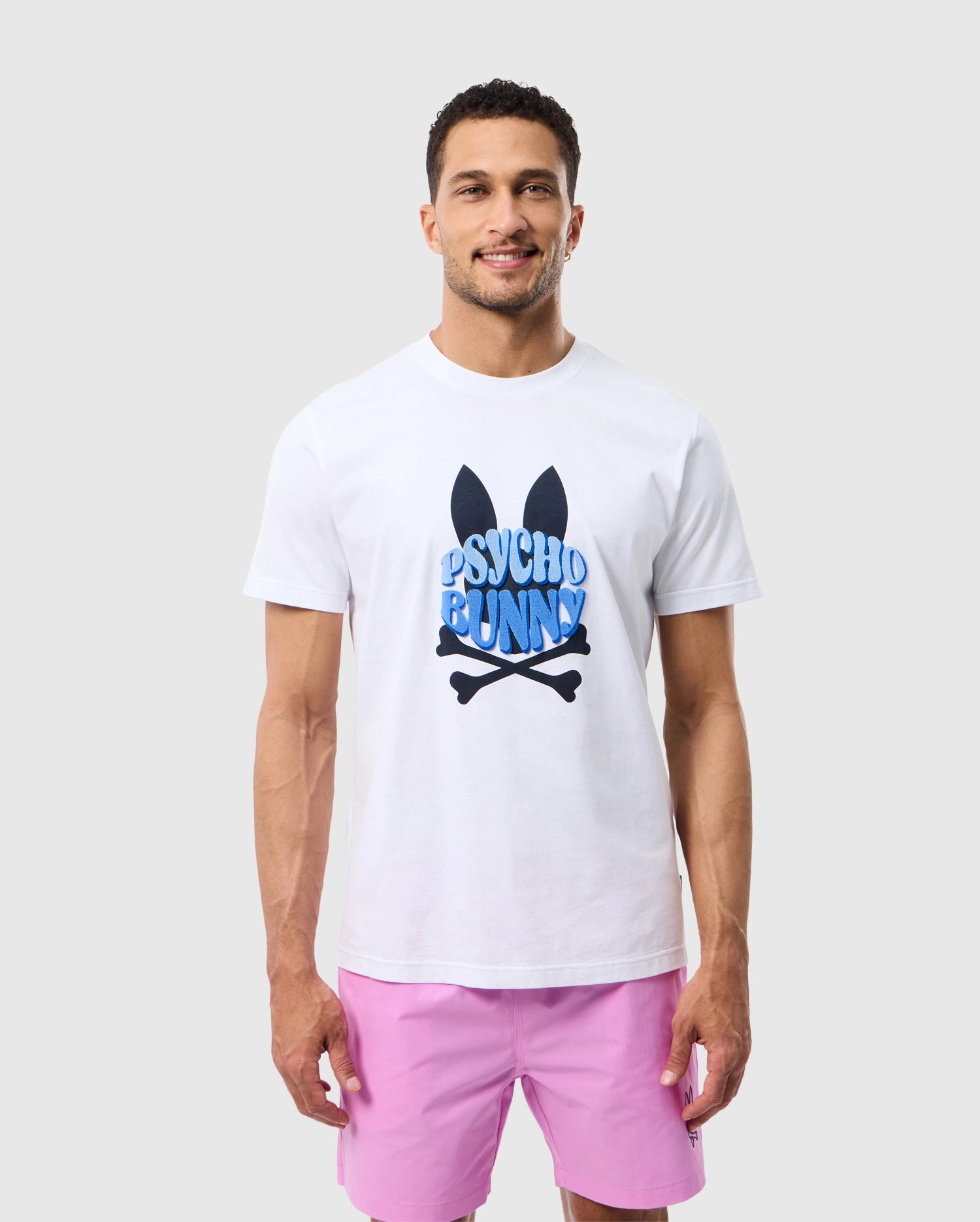 A man stands against a plain background, smiling at the camera while wearing the MENS PRESTON GRAPHIC TEE - B6U611C200 from Psycho Bunny. The Pima cotton t-shirt features a playful 