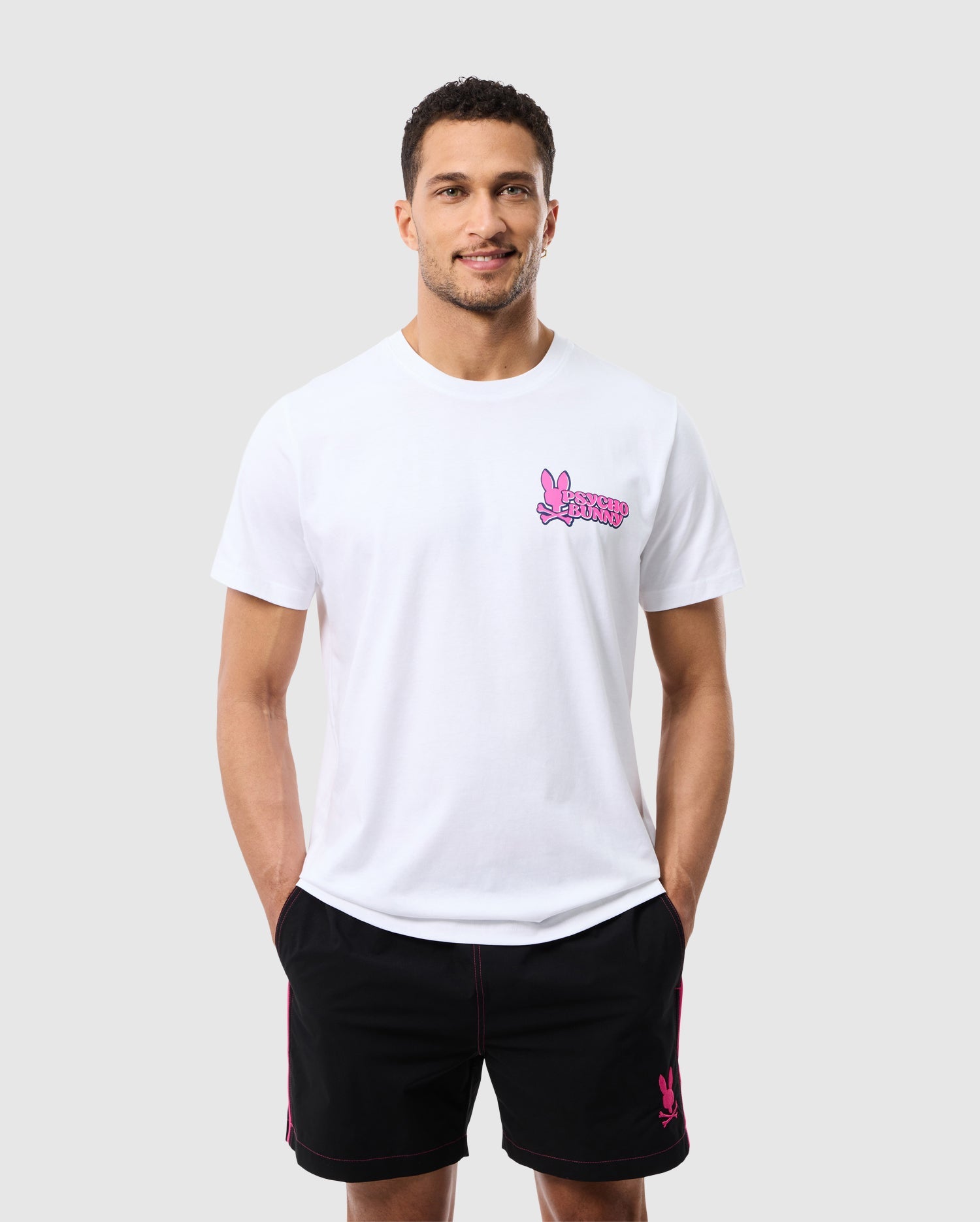 A smiling man with short dark hair is wearing the MENS REDLAND GRAPHIC TEE - B6U613C200 by Psycho Bunny, which is a white T-shirt featuring a pink logo with a peace hand sign that evokes a tropical aesthetic. He also has on black shorts with pink accents. His hands are in his pockets, and the background is plain and light gray.