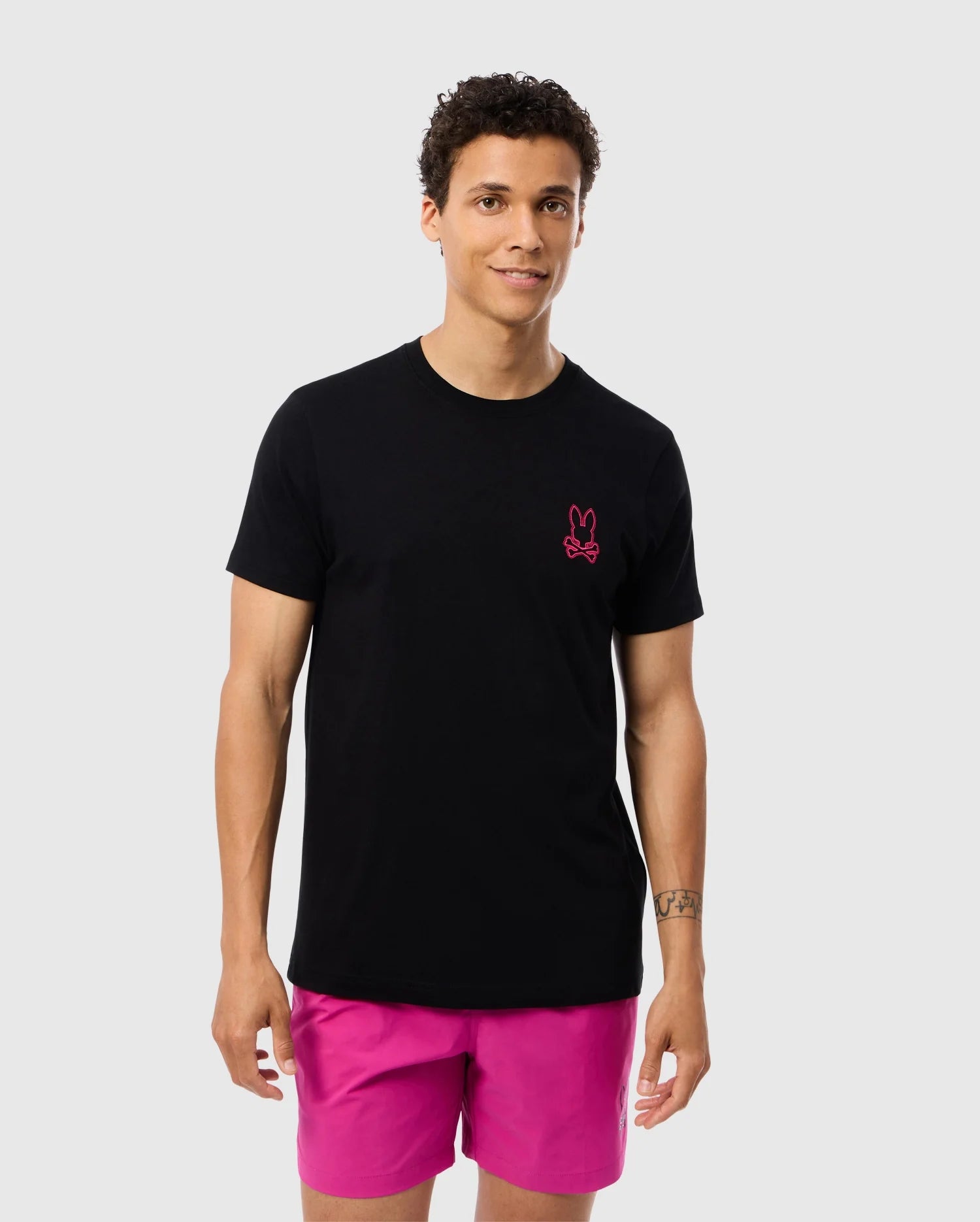 A person with short curly hair wearing a black Psycho Bunny Pima cotton tee (MENS PEARLAND BACK GRAPHIC TEE - B6U632C200) featuring a small pink bunny logo on the left chest and bright pink shorts stands against a plain light grey background. They are looking forward with a slight smile.