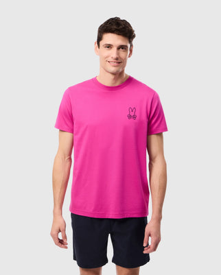 A man with short dark hair is standing and smiling at the camera. He is wearing a bright pink Psycho Bunny MENS PEARLAND BACK GRAPHIC TEE - B6U632C200 made of soft Pima cotton, featuring a small bunny graphic on the left side of the chest, paired with dark blue shorts. The background is plain light grey.