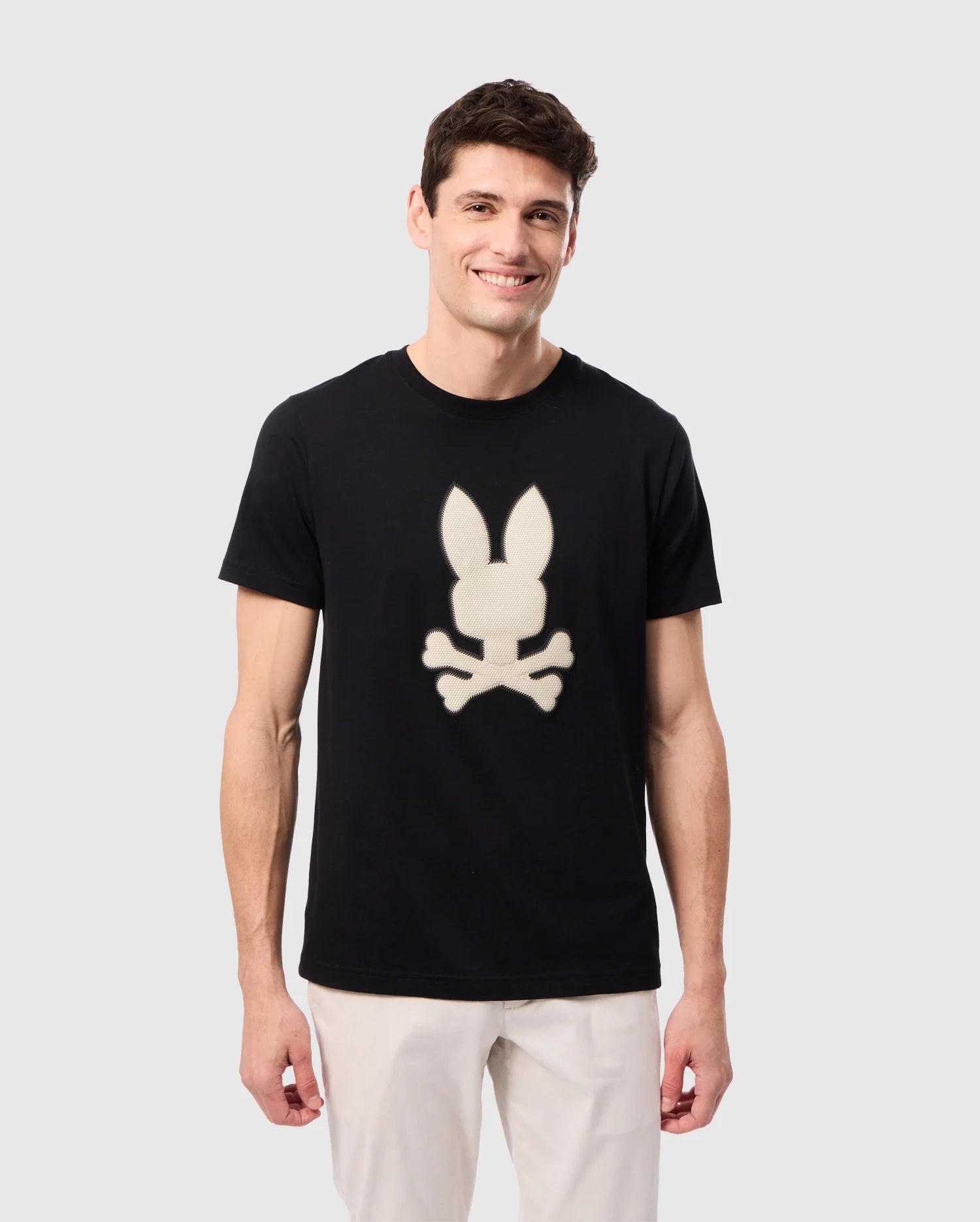 A person with short dark hair is smiling while wearing the Psycho Bunny MENS RIVIERA GRAPHIC TEE - B6U639C200, made of soft Peruvian Pima cotton, featuring a white bunny head with ears up and crossbones below. The person is also dressed in light-colored pants and standing against a plain light grey background.