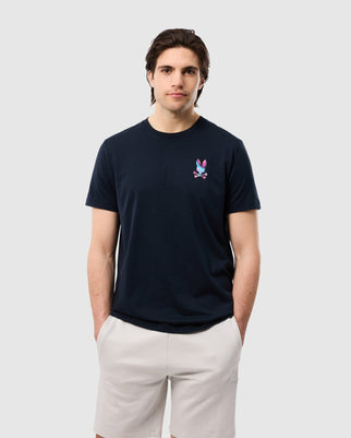 A man with dark hair is wearing a black MENS TYLER BACK GRAPHIC TEE - B6U674C200 from Psycho Bunny made from 100% Pima cotton, featuring a colorful, small bunny print logo on the left side of the chest. He is also wearing light-colored shorts and has his hands in his pockets. He stands against a plain, light grey background.