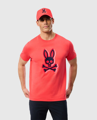 A person dressed in the Psycho Bunny MENS KAYDEN GRAPHIC TEE - B6U676C200, a bright red Pima cotton t-shirt adorned with a dark bunny skull and crossbones design at the center. They complement the look with black pants and a matching red cap, both embroidered with the same distinctive bunny skull logo. The background is plain white.