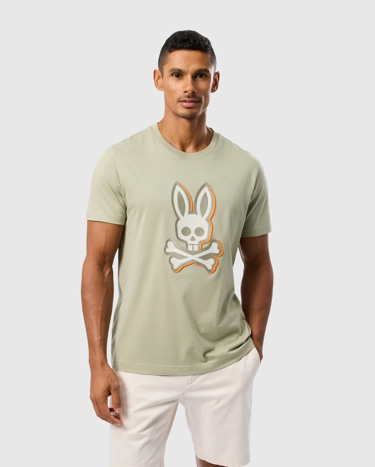 A man with short hair is wearing a light green Psycho Bunny MENS KAYDEN GRAPHIC TEE - B6U676C200 featuring an embroidered Bunny skull and crossbones, paired with white shorts. He is standing against a plain, light gray background.