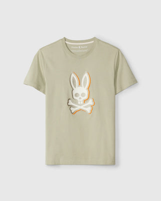 A light beige Pima cotton graphic tee with a white and gray skull and crossbones rabbit logo on the front. The skull has rabbit ears, and the crossbones are placed underneath. This Regular fit t-shirt features "Psycho Bunny" text on the collar with a small embroidered bunny logo above it. This is the Psycho Bunny MENS KAYDEN GRAPHIC TEE - B6U676C200.