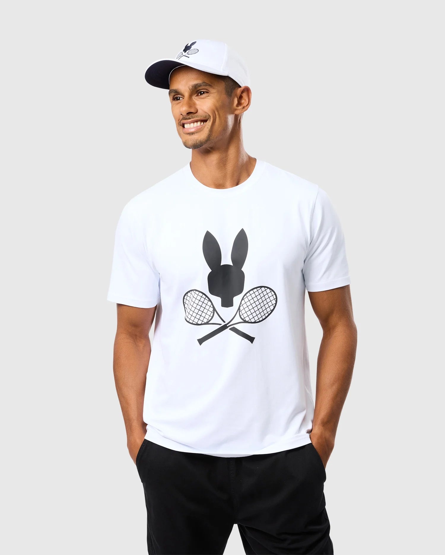 A man, dressed in a Psycho Bunny MENS COURTSIDE GRAPHIC TEE - B6U681C200, showcases a bunny silhouette over two crossed tennis rackets. He complements his outfit with a white cap and black pants, smiling with his hands in his pockets against a plain background.