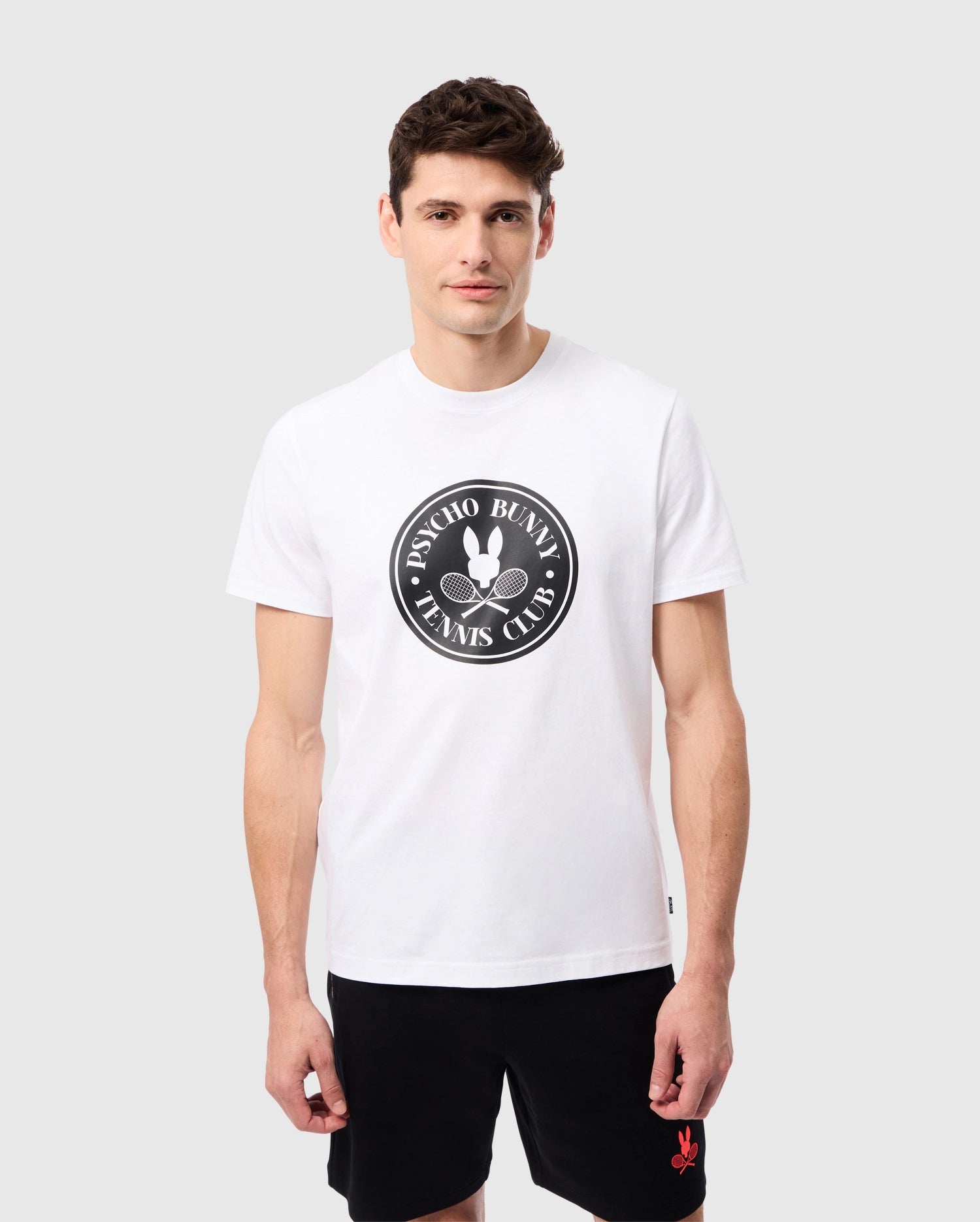 A man stands against a plain light gray background wearing the Psycho Bunny Men's Courtside Graphic Tee, which is made from soft Pima cotton jersey and features a 