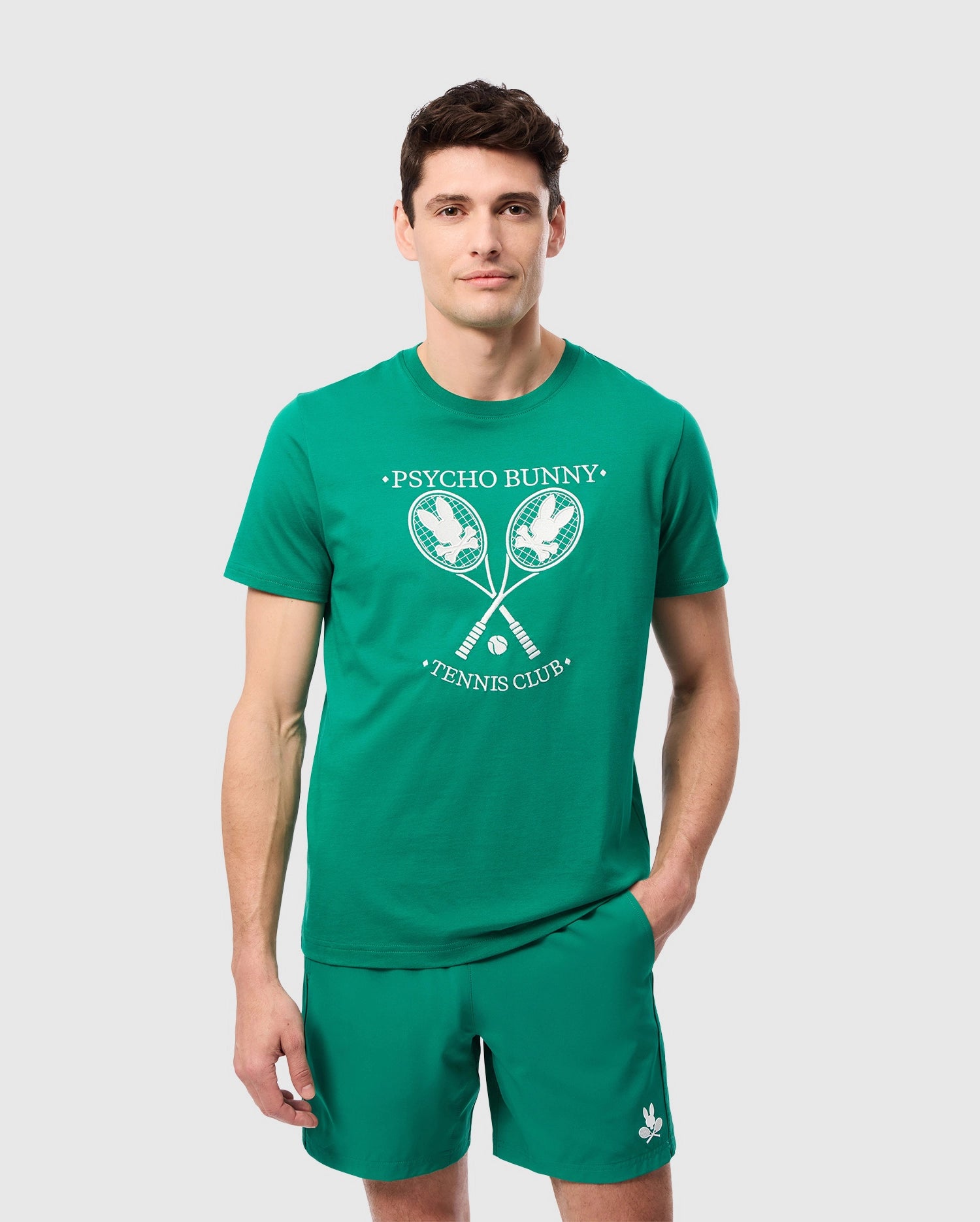 A man stands against a plain background wearing a green Pima cotton jersey t-shirt and matching green shorts. The Psycho Bunny MENS COURTSIDE GRAPHIC TEE features two crossed tennis rackets and the words 