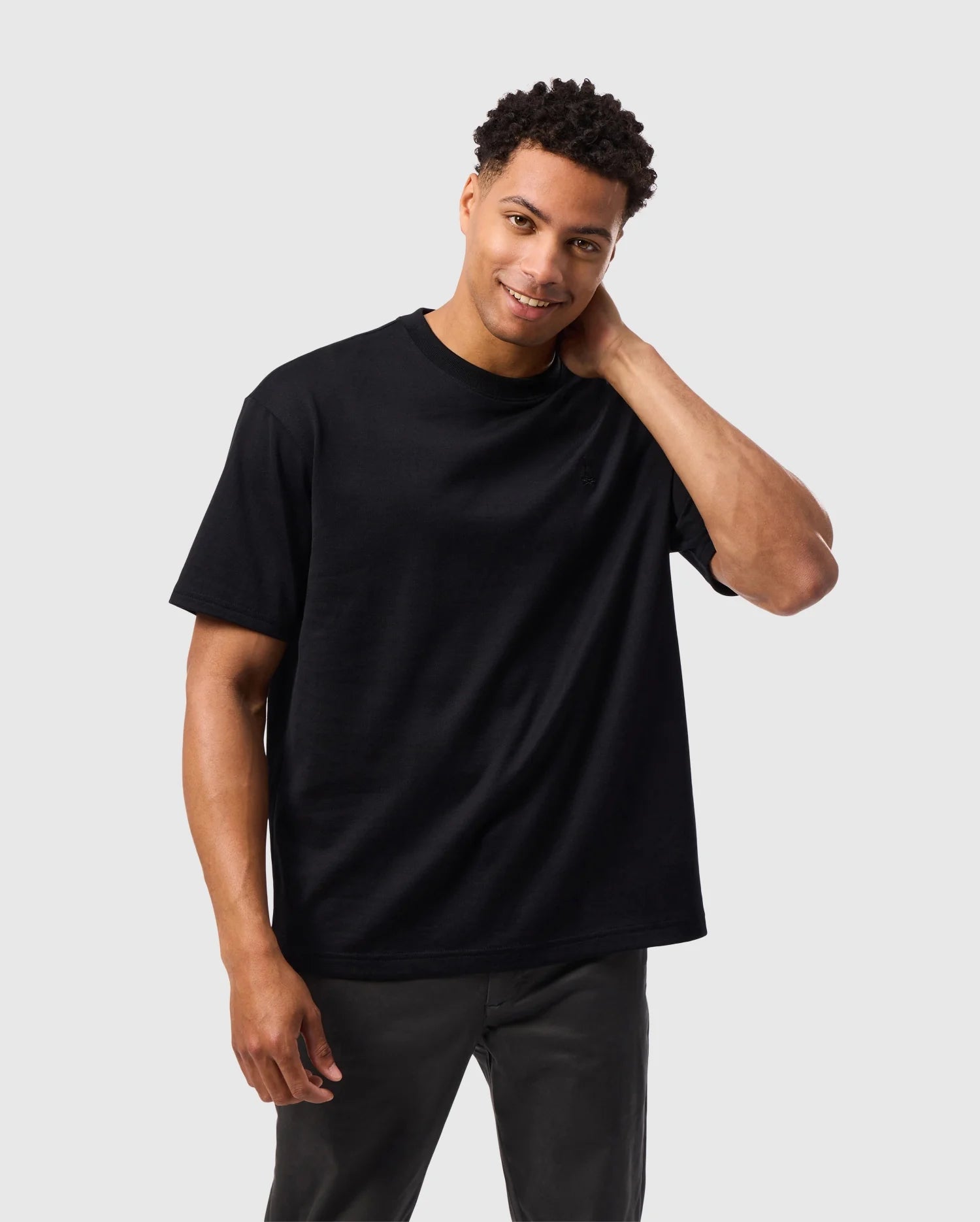 A man with short, curly hair is smiling and touching his neck with his right hand. He is wearing a Psycho Bunny MENS DUKE OVERSIZED TEE - B6U742D200 in black, made of heavyweight Pima cotton, paired with dark-colored pants against a plain, light gray background.