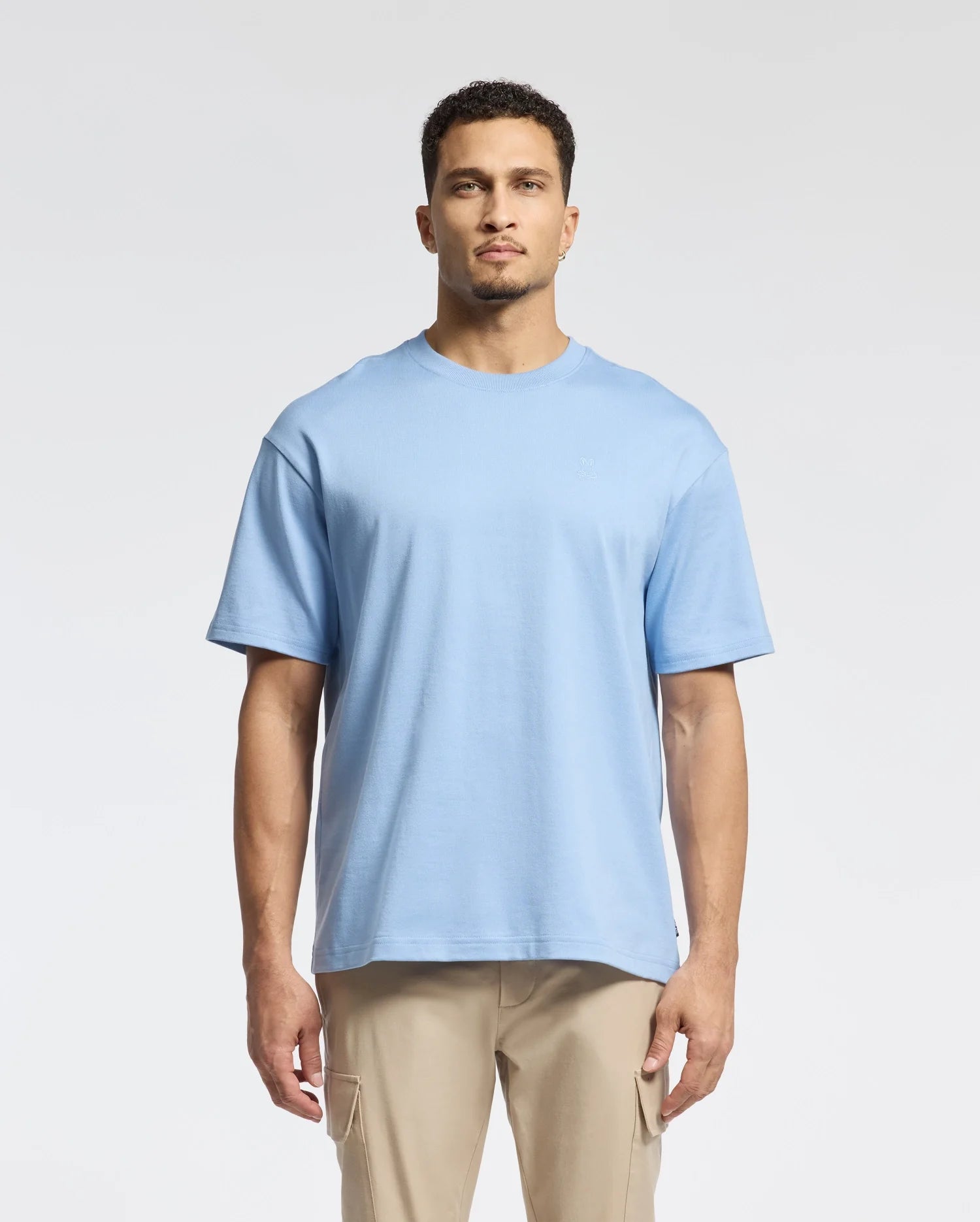 A man stands wearing a light blue tee and beige pants against a plain white background. The T-shirt is the Psycho Bunny MENS DUKE HEAVY WEIGHT RELAXED FIT TEE - B6U742D200, crafted from heavyweight jersey with a simple design.