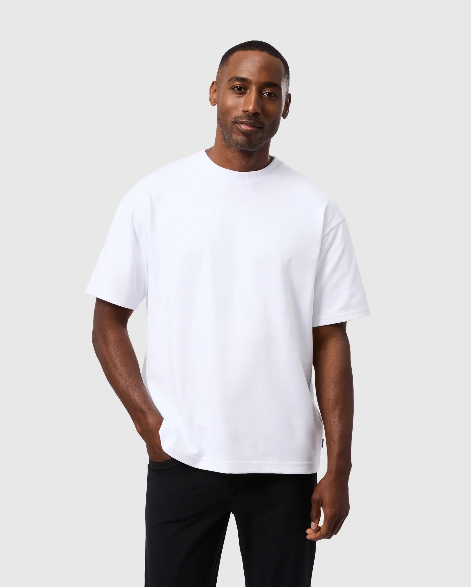 A man with short hair, wearing a Psycho Bunny MENS DUKE OVERSIZED TEE - B6U742D200 made of heavyweight Pima cotton and black pants, stands against a plain light gray background. He is looking at the camera with a relaxed posture, one hand in his pocket, and a slight smile on his face.