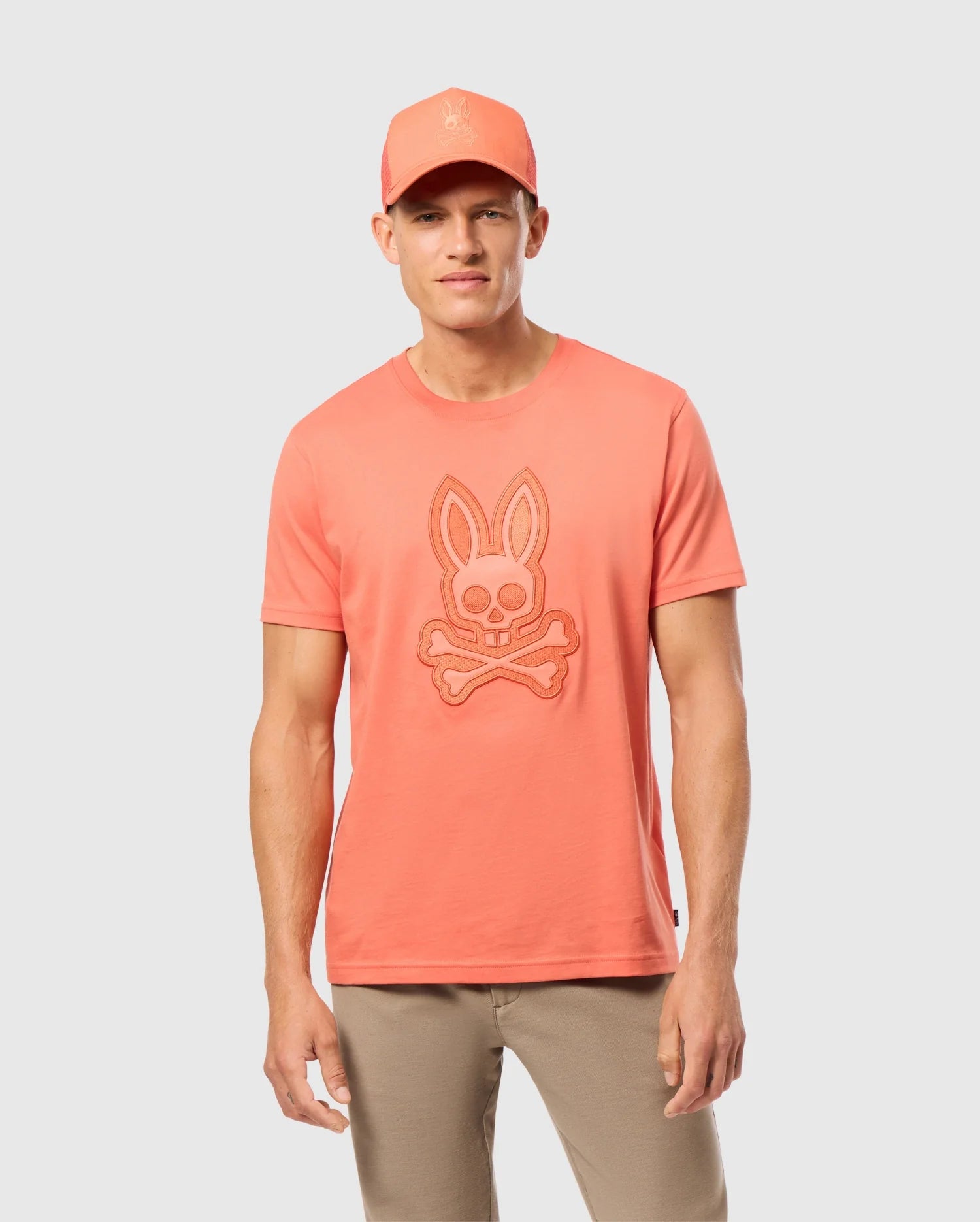 Against a light gray background, a man stylishly wears the Psycho Bunny MENS MALTON GRAPHIC TEE - B6U795D200 in coral, made from soft Pima cotton with a bunny skull and crossbones logo. He completes the look with a matching coral baseball cap and beige pants.