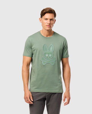 A man with short brown hair wears a green Psycho Bunny MENS MALTON GRAPHIC TEE - B6U795D200, featuring a bunny skull and crossbones design on the front. He is also wearing dark gray pants and standing against a plain light gray background.