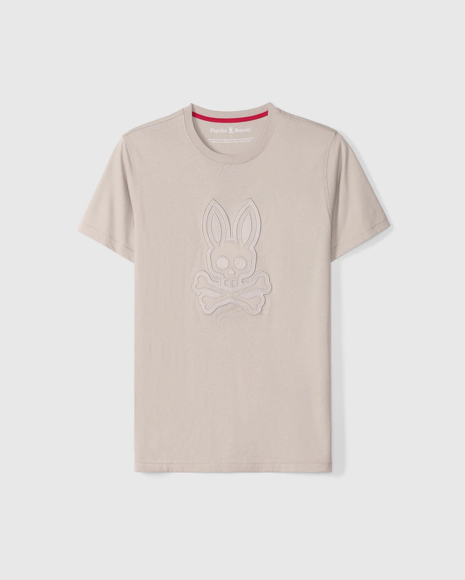 The Psycho Bunny MENS MALTON GRAPHIC TEE - B6U795D200 is a beige graphic tee featuring a rabbit skull and crossbones design on the front, outlined in stitched patterns of the same color. Crafted in Peru from soft Pima cotton fabric, it includes a contrasting red inner band at the neckline with white text that reads 