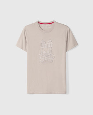 The Psycho Bunny MENS MALTON GRAPHIC TEE - B6U795D200 is a beige graphic tee featuring a rabbit skull and crossbones design on the front, outlined in stitched patterns of the same color. Crafted in Peru from soft Pima cotton fabric, it includes a contrasting red inner band at the neckline with white text that reads "Psycho Bunny.