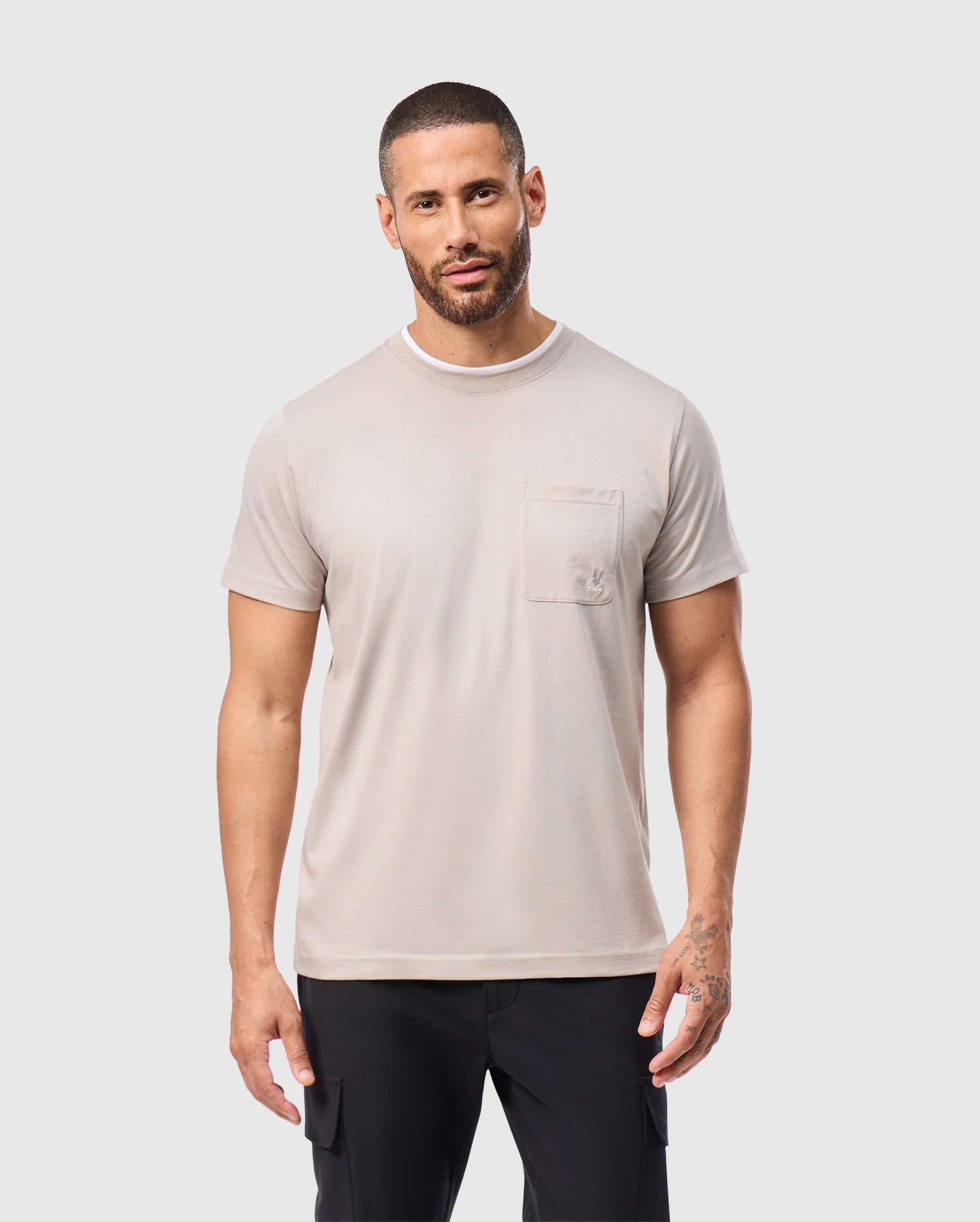 A bearded man stands against a plain white background, dressed in a soft pima cotton MENS CAMPO POCKET TEE by Psycho Bunny in light beige, paired with black pants. His short hair and tattoos on his left hand enhance his distinctive appearance.