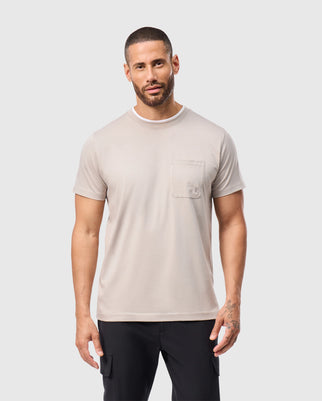 A bearded man stands against a plain white background, dressed in a soft pima cotton MENS CAMPO POCKET TEE by Psycho Bunny in light beige, paired with black pants. His short hair and tattoos on his left hand enhance his distinctive appearance.