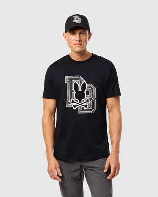 A man wearing a black Psycho Bunny Men's Zack Graphic Tee (B6U897D200) with a large white outlined "D" and "B" logo featuring a silhouette of a bunny head with crossed bones below it is standing against a plain light gray background.