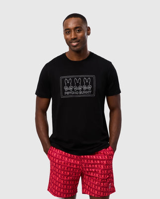 A man stands against a light gray background, wearing the MENS TACOMA GRAPHIC TEE - B6U909D200 from Psycho Bunny, which is made of black Pima cotton and features a print of three bunny heads along with the words "Psycho Bunny." He is also wearing red patterned shorts and has his hands casually in his pockets. He is smiling and looking at the camera.