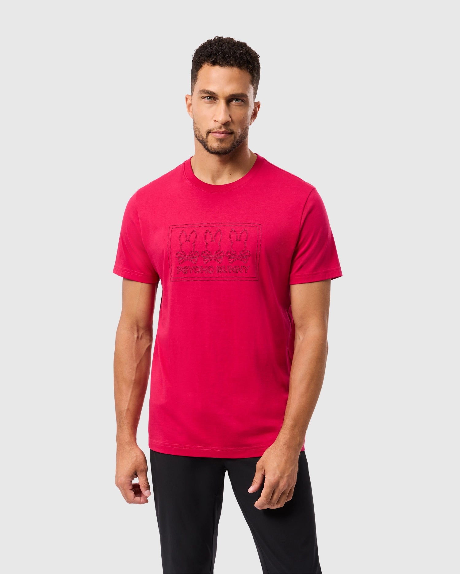 A man with short curly hair and a trimmed beard is wearing a bright red Psycho Bunny Men's Tacoma Graphic Tee (B6U909D200) made from soft Pima cotton, featuring multiple cartoon bunnies and the text 