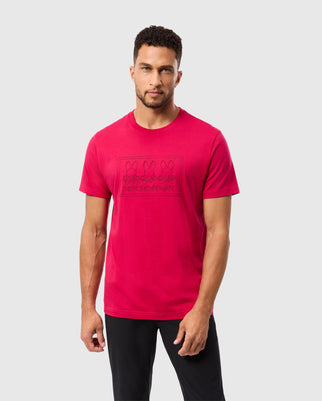 A man with short curly hair and a trimmed beard is wearing a bright red Psycho Bunny Men's Tacoma Graphic Tee (B6U909D200) made from soft Pima cotton, featuring multiple cartoon bunnies and the text "Beyond Bunny." He is standing against a plain light gray background, looking straight ahead with a neutral expression.