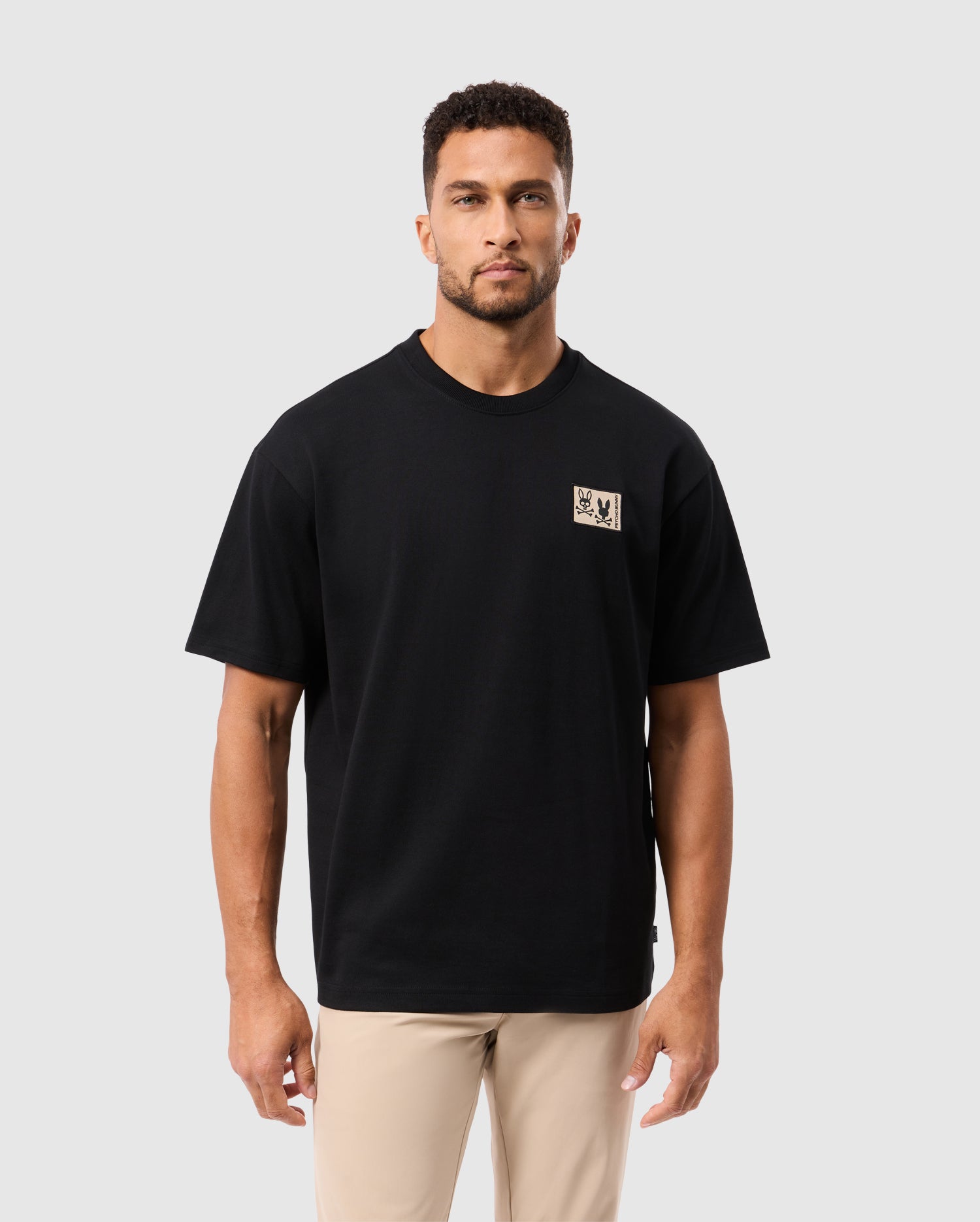 Wearing a Psycho Bunny MENS MONROE HEAVY WEIGHT RELAXED FIT TEE - B6U910D200 in black, made from premium Pima cotton with a stylized chest logo patch, this person enjoys the comfortable drape as they stand against a plain white background.