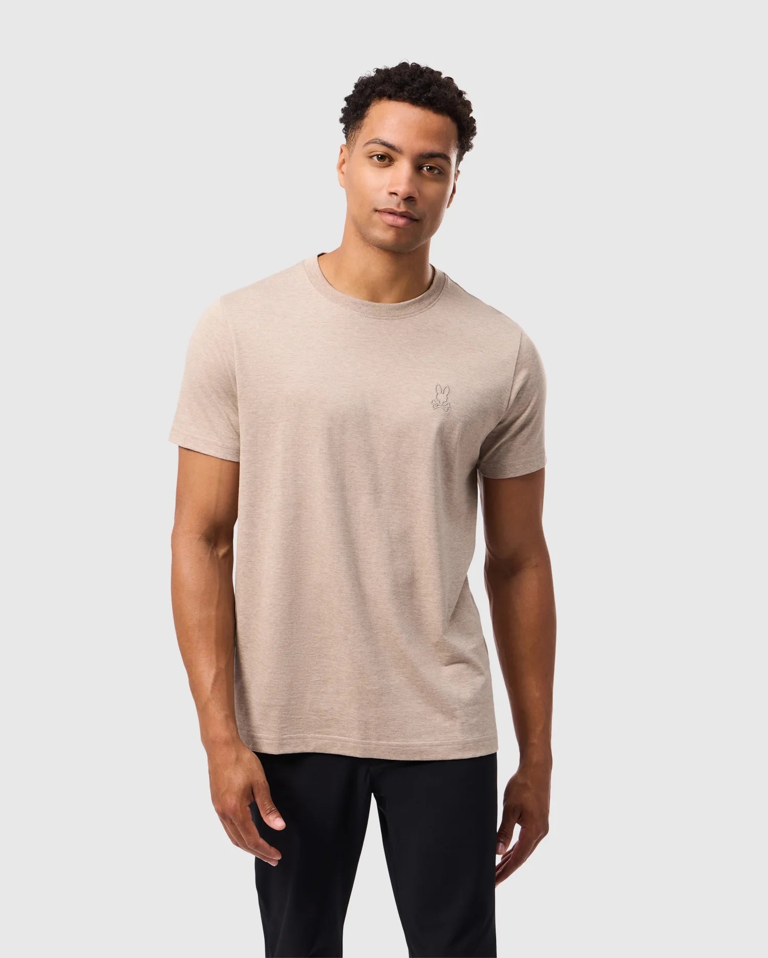 A person with short curly hair stands against a plain light gray background, wearing a beige Psycho Bunny MENS JAMES PREMIUM CREW NECK TEE made from premium Pima cotton and black pants. The t-shirt, model B6U952D200, features meticulous embroidery on the left chest. The person has a relaxed, neutral expression and is facing the camera.