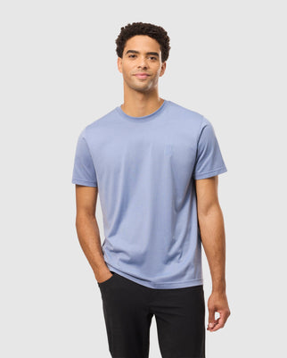 A man with short, curly hair is wearing the Psycho Bunny MENS JAMES PREMIUM CREW NECK TEE (B6U952D200) in light blue, made from premium Pima cotton, along with black pants. He stands in a regular fit style with one hand in his pocket, smiling slightly against a plain white background.