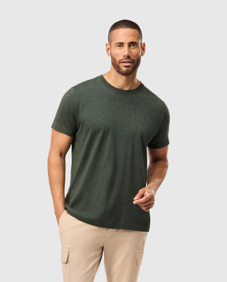 A man with short hair and a beard is wearing a dark green, stylish Psycho Bunny MENS JAMES PREMIUM CREW NECK TEE - B6U952D200 and beige cargo pants. He is standing against a light grey background with his right hand partially in his pocket and left hand slightly raised.