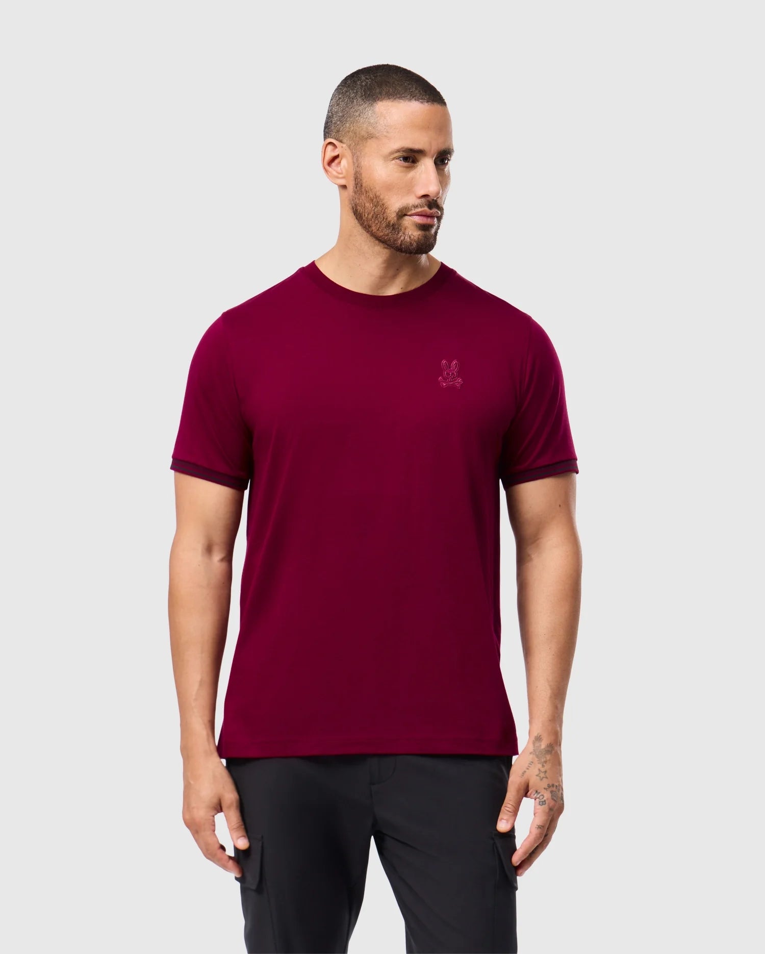 A man with short hair and a trimmed beard is wearing a maroon MENS MALTON FASHION TEE - B6U966D200 by Psycho Bunny and black pants. He stands against a plain grey background with his arms at his sides. A small embroidered logo can be seen on the left chest of the stylish cotton t-shirt.