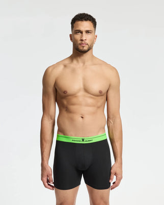 A man stands with a neutral expression, wearing Psycho Bunny's MENS 3 PACK BOXER BRIEF - B6V347E200 in black featuring a green waistband. Made from luxurious Pima cotton, they guarantee comfort. The room is brightly lit, and the background is plain white.