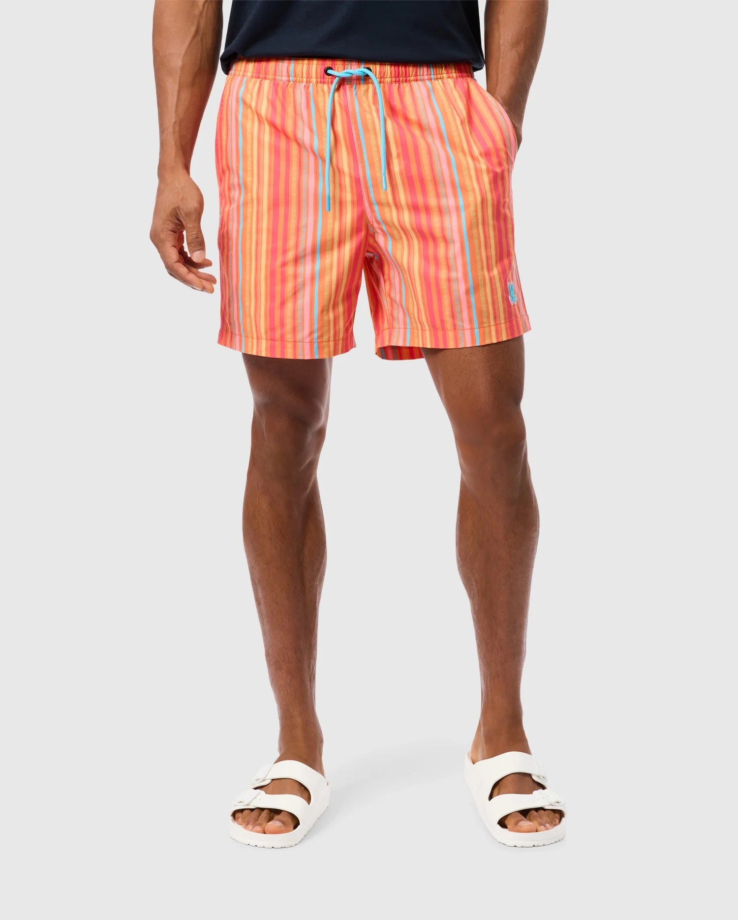 A person is standing against a plain background, showcasing vibrant Psycho Bunny MENS ANTON ALL OVER PRINT SWIM TRUNK - B6W542C200 in hues of pink, red, and orange, secured by a blue drawstring. They are also wearing white sandals. The photo captures the body from mid-torso down to the feet.