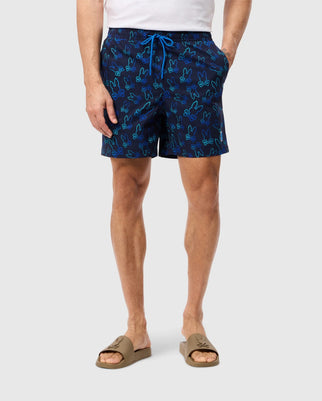 A person dressed in a white shirt and navy blue Psycho Bunny MENS SHELDON ALL OVER PRINT SWIM TRUNK - B6W588C200, showcasing a comprehensive Bunny pattern, stands against a plain background. They are also wearing tan slide sandals.