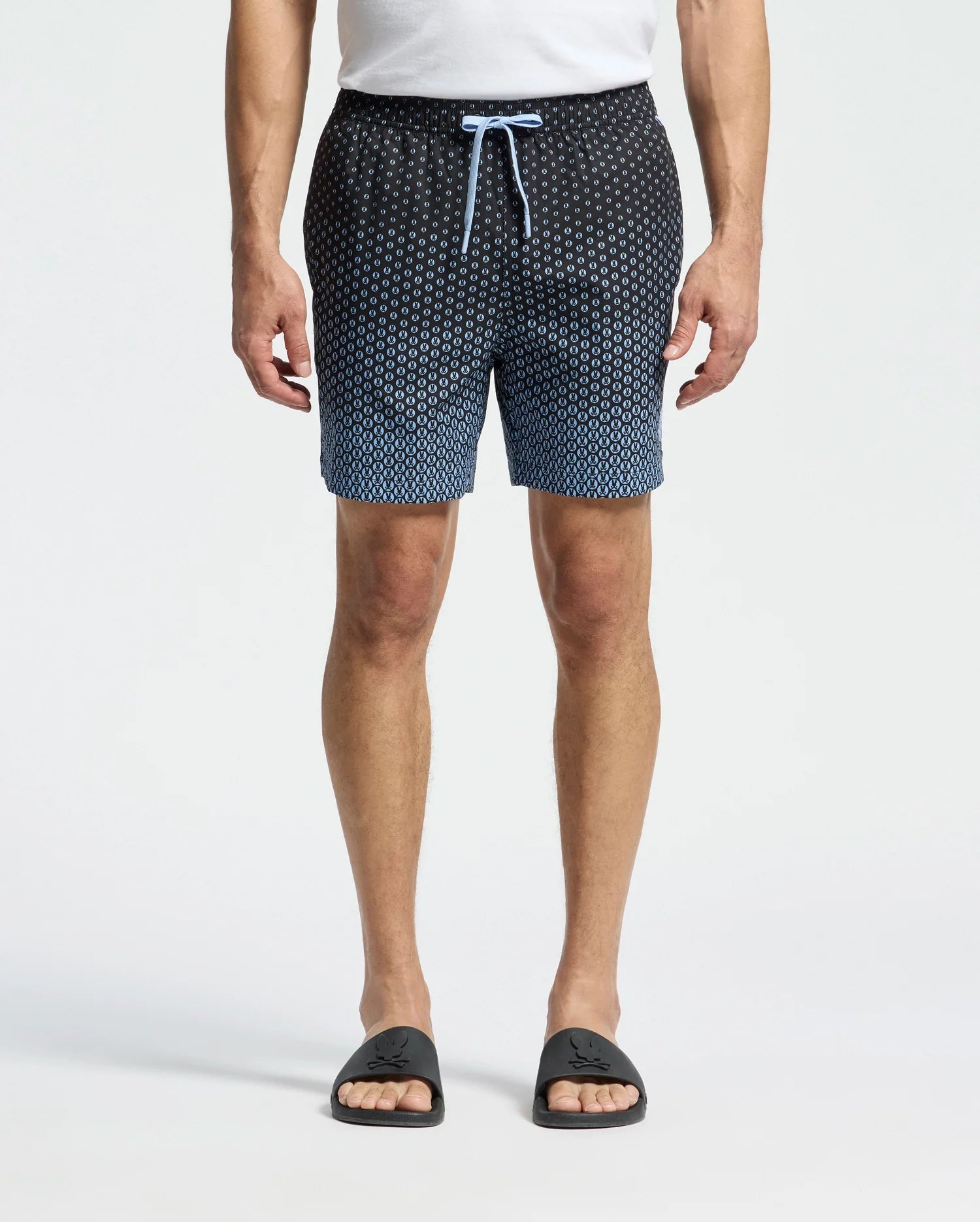 A person wears Psycho Bunny's MENS BENDIGO ALL OVER PRINTED SWIM TRUNK - B6W619F200, featuring a black to blue gradient with light blue drawstrings, facing forward. They pair it with logo-adorned black slide sandals and have a waterproof bag beside them against a plain, light-colored backdrop.