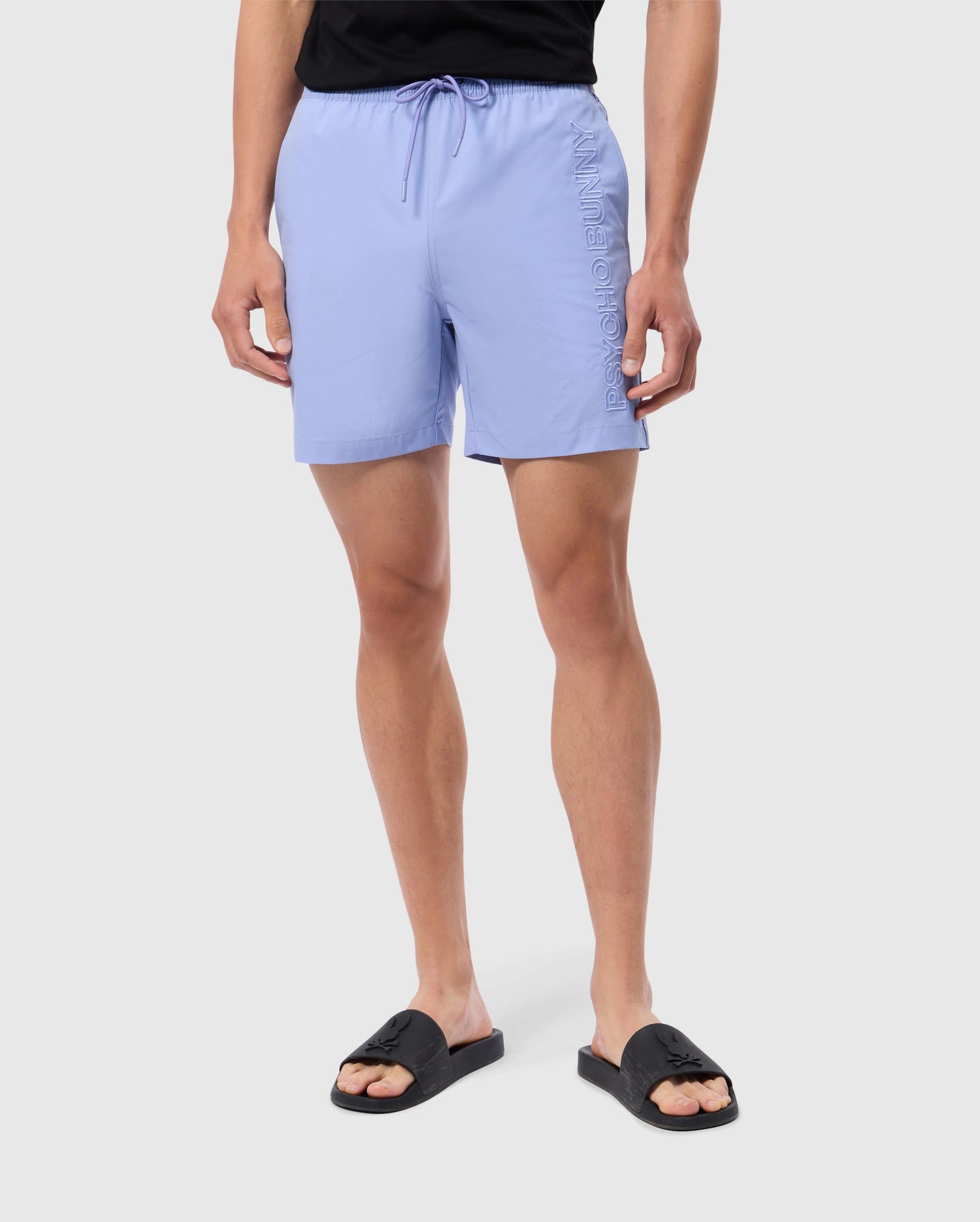 A person is sporting Psycho Bunny's light blue MENS MELVIN SWIM TRUNK with a drawstring and quick-dry fabric, paired with black slide sandals. The swim trunk showcases text embossed vertically on the side against a plain gray background.