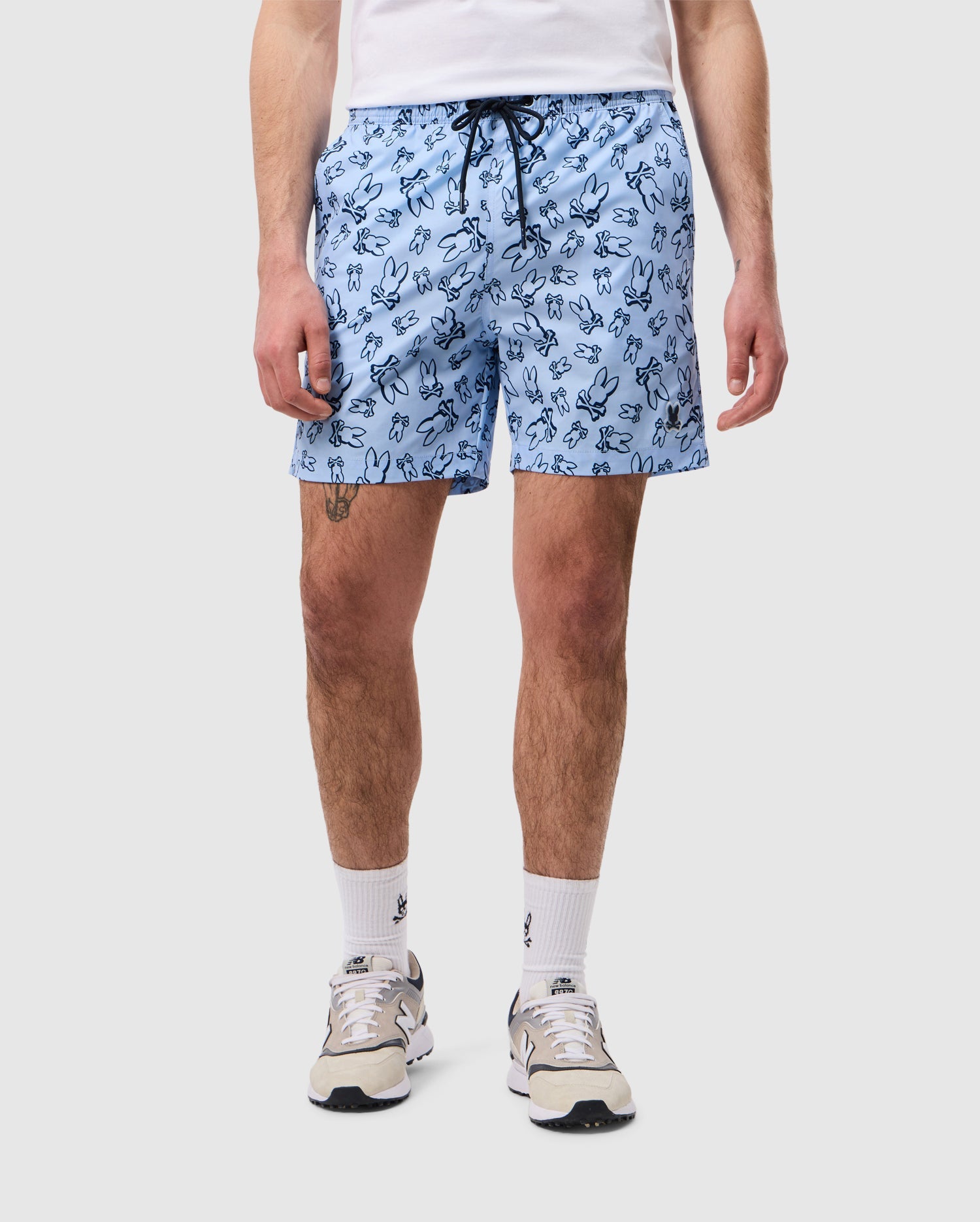 A person is standing against a plain background, wearing a white T-shirt, Psycho Bunny's light blue MENS RENDON PRINT SWIM TRUNK - B6W669C200 featuring a black cartoon bunny print, white socks with a small black logo, and white sneakers with gray and beige accents. The person's face is not visible in the image.