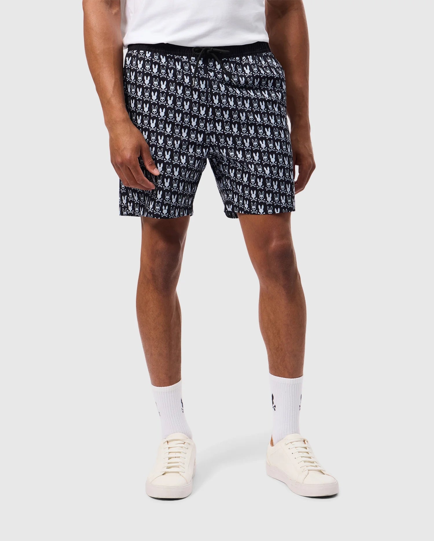 A person wearing Psycho Bunny's MENS TILDEN PRINT SWIM TRUNK - B6W729D200, which features a repetitive, contrasting bunny print in black and white, stands against a plain background. They are paired with white socks and white sneakers. The person’s upper body is not visible.