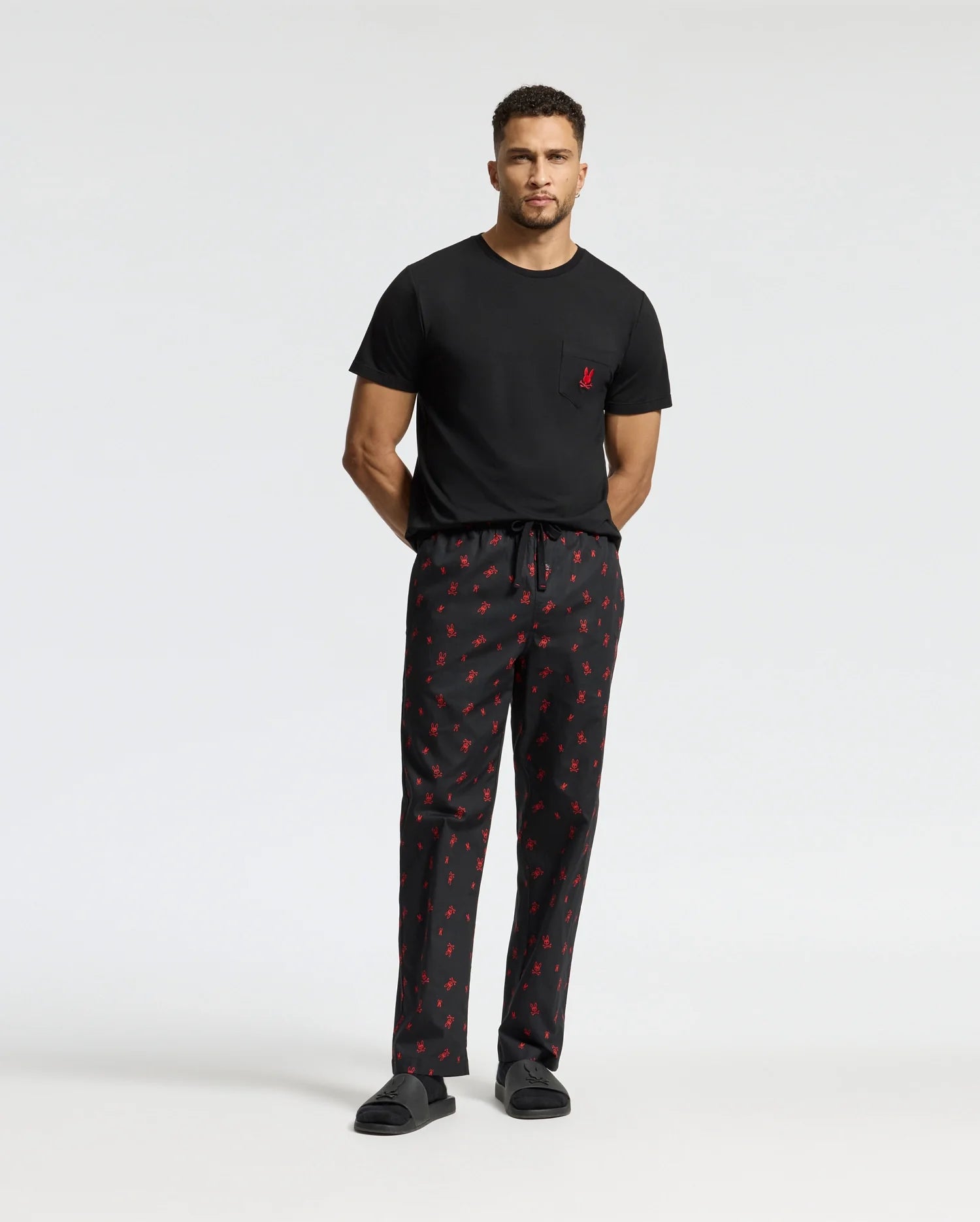 A man with short curly hair stands against a plain background wearing the Psycho Bunny MENS TELRIDE PRINTED PAJAMA SET, featuring a comfortable black T-shirt and pajama pants adorned with red patterns. He completes his cozy sleepwear ensemble with black slippers, his hands positioned behind his back.