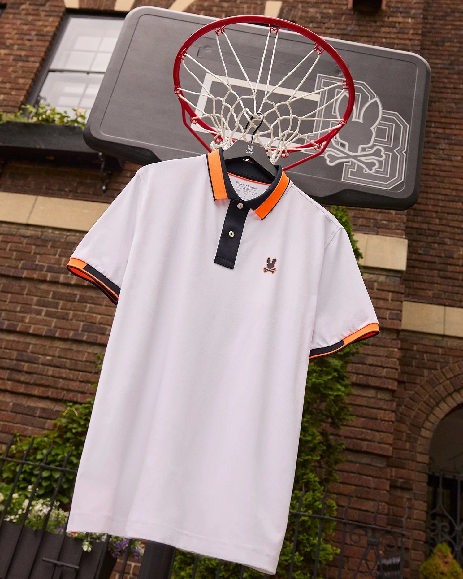 A Psycho Bunny MENS BELLEVUE SPORT POLO SHIRT - B6K905D200 made of micro mesh fabric with orange and black trim hangs from a black basketball hoop's net outside a brick building. The shirt, featuring a small embroidered logo on the chest, is attached to a rectangular backboard boasting an intricate design.